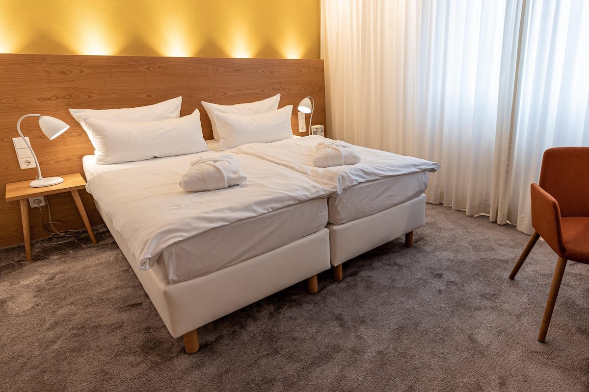 Quarto de hotel com uma cama grande, duas luminárias de cabeceira, uma cabeceira de madeira, roupa de cama branca e um tapete castanho.
