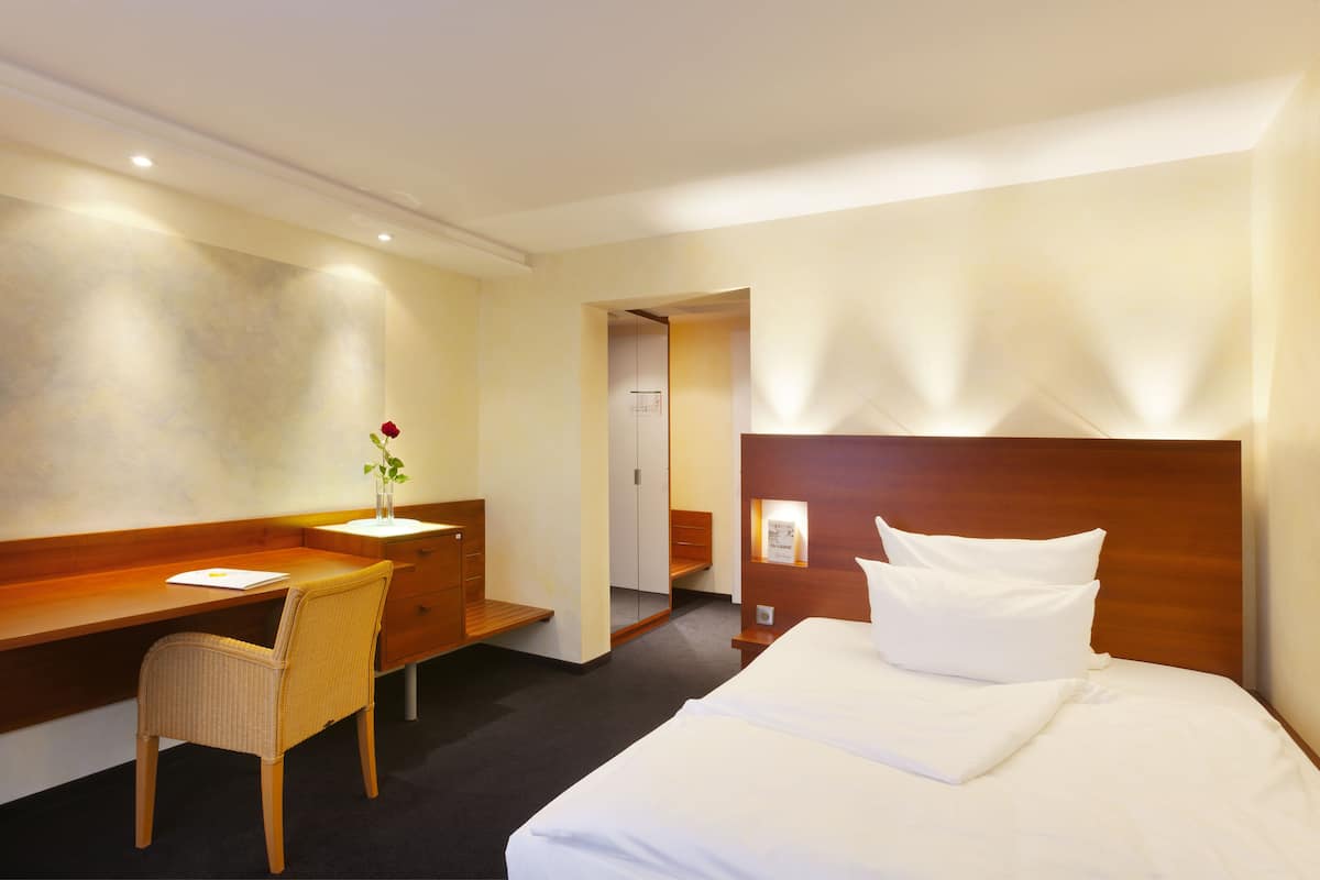 Quarto de hotel com escrivaninha de madeira, uma cadeira, cama com roupa branca e cabeceira de madeira.