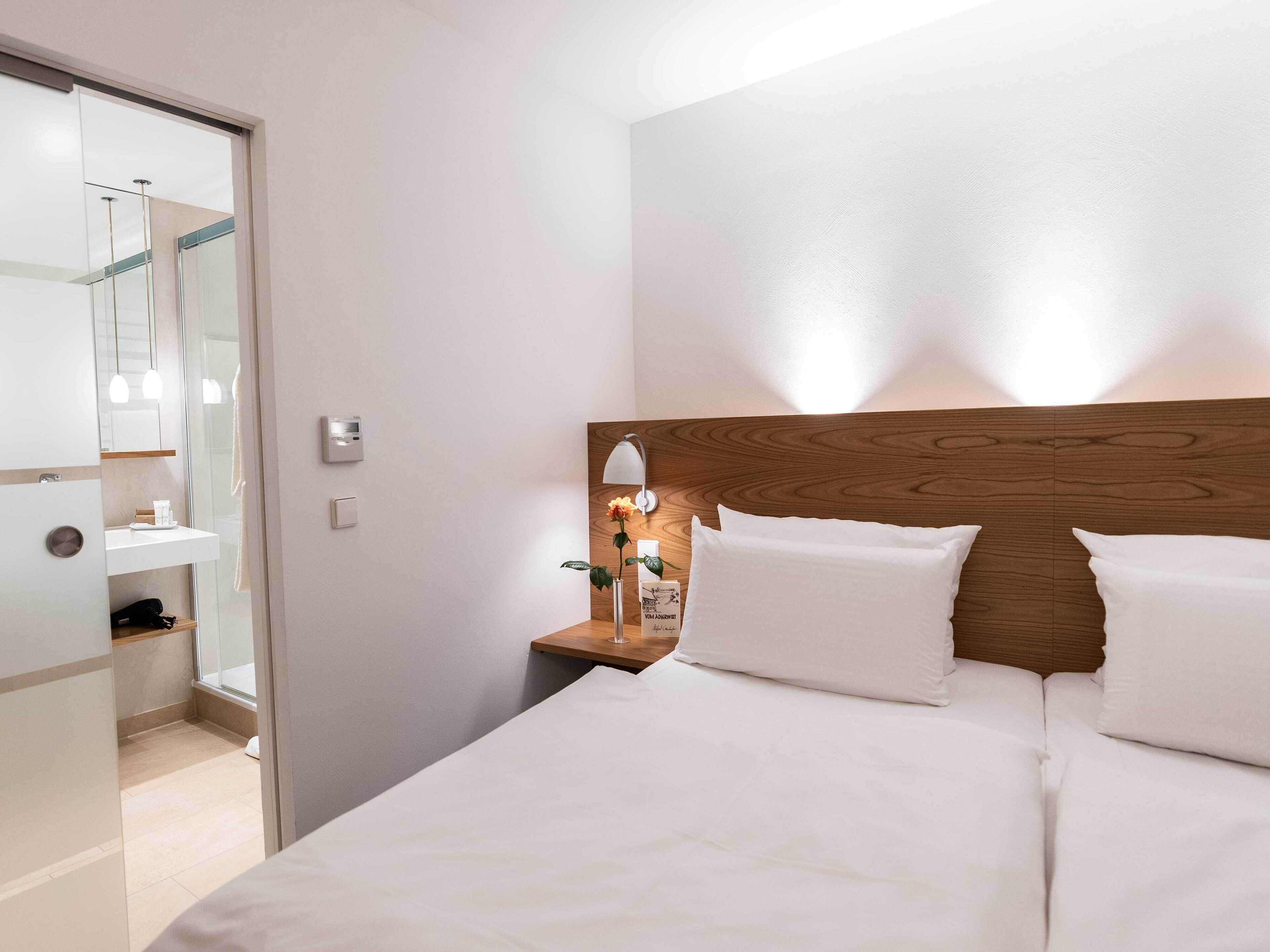 A hotel room with a wooden headboard, a bedside table with a lamp, and a large bed with white bedding.