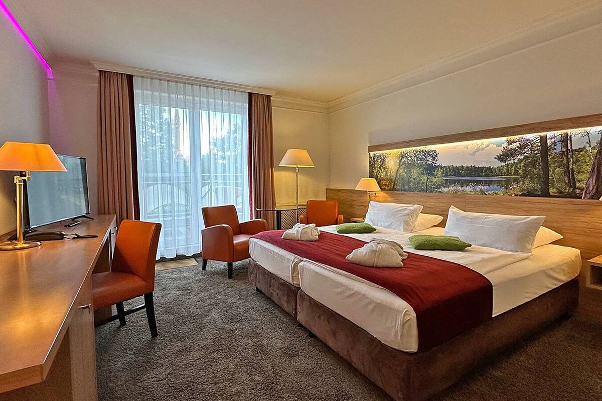A hotel room with a bed, two chairs, a desk, a TV, and a window with a view of trees.
