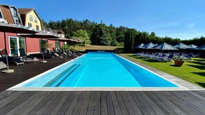 A rectangular swimming pool with a wooden deck surrounding it. There are lounge chairs and umbrellas on the right side. - Hotel & Spa Sommerfeld - Adults Only (Kremmen)