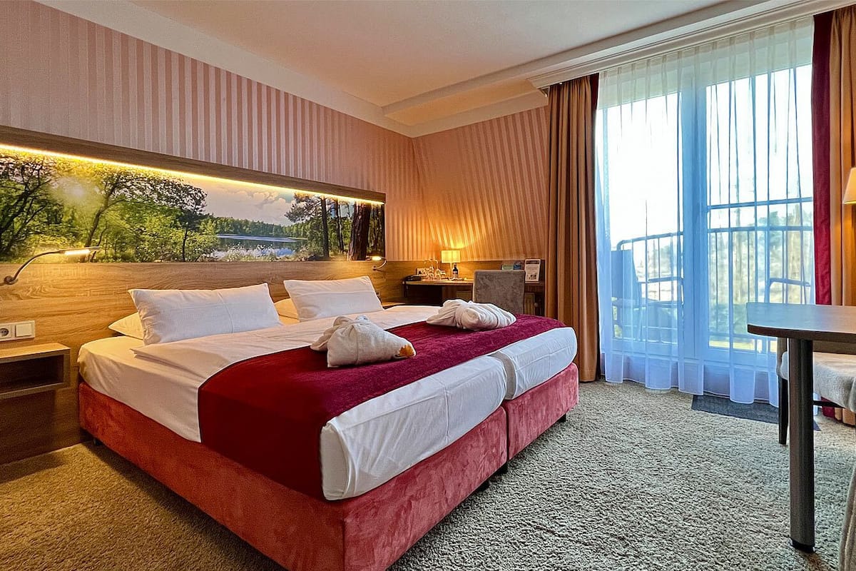 A hotel room with a large bed, a desk, a chair, a nightstand, and a wall-mounted lamp.