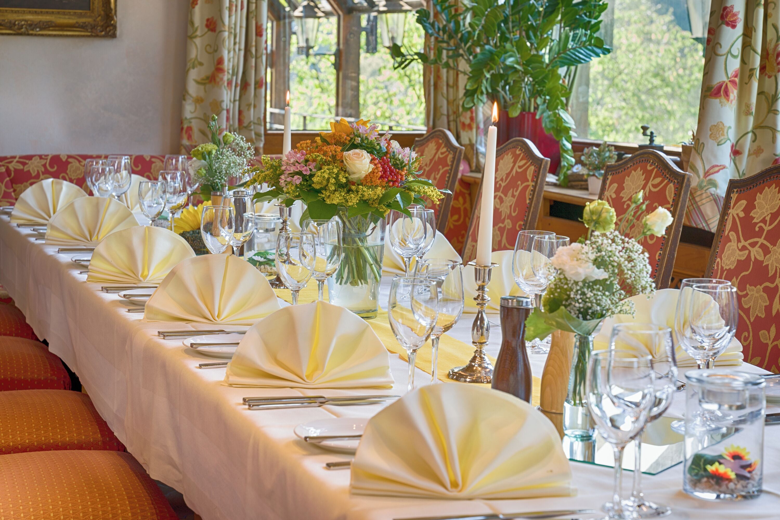 Een lange eettafel gedekt met witte tafelkleden, gele servetten en glazen, omringd door rode fauteuils.