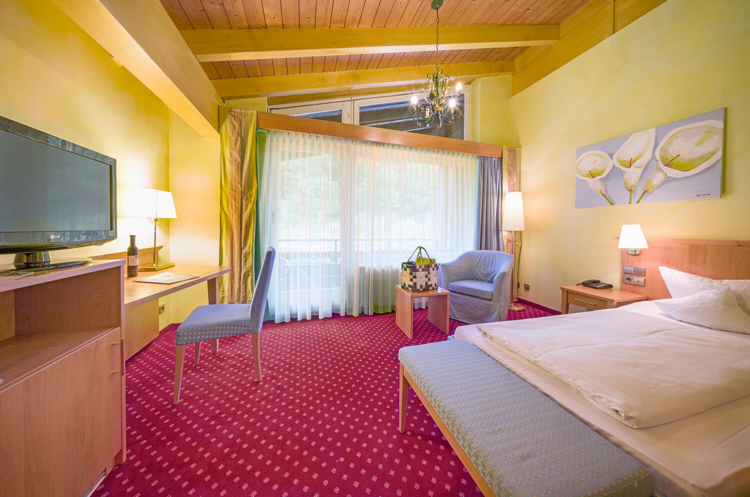 Quarto de hotel com uma cama, uma escrivaninha com televisão, uma cadeira, uma mesinha de cabeceira com abajur e uma janela com cortinas.