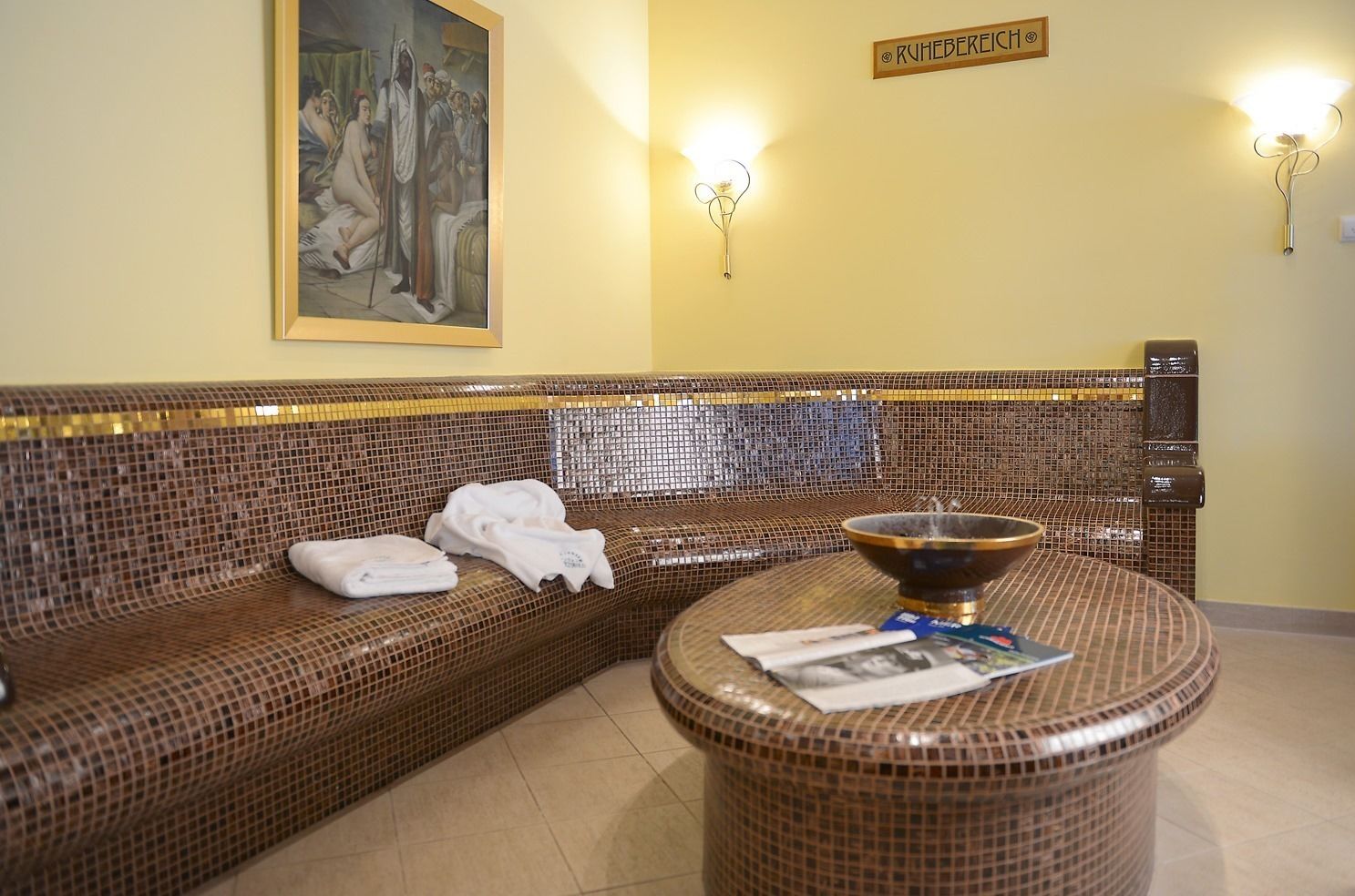A room with a mosaic-patterned sofa, a round table with a bowl and magazines, and a painting on the wall.