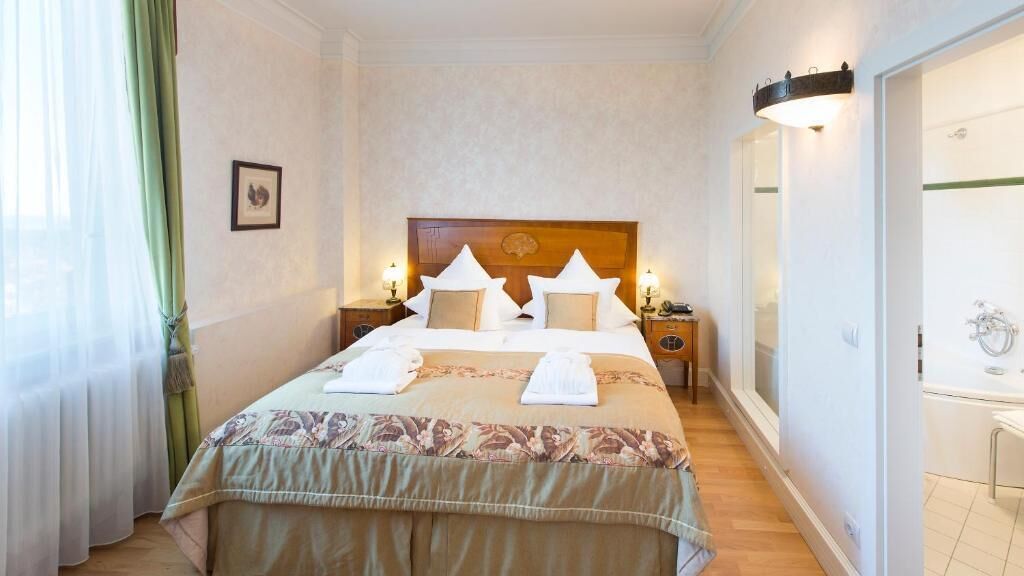 A hotel room with a large bed, a wooden headboard, bedside lamps, a nightstand, and a bathroom with a mirror and sink.