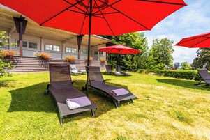 A patio area with two reclining chairs, each under a red umbrella, and a building with a porch in the background.