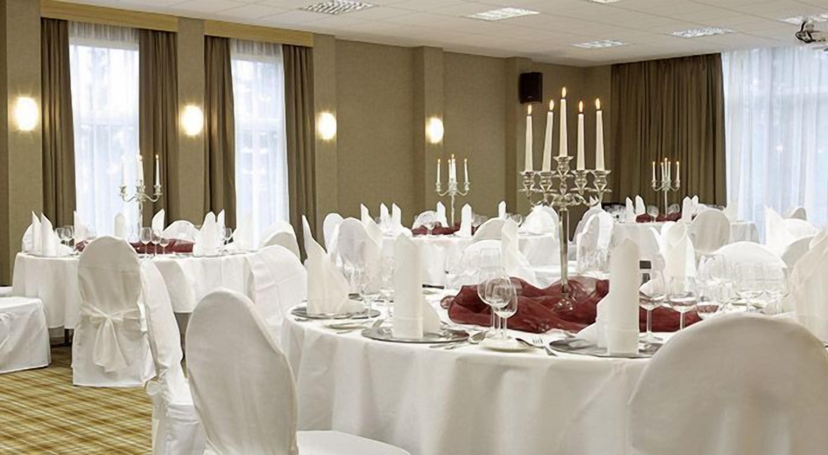 Ein Festsaal mit runden Tischen, gedeckt für eine festliche Veranstaltung, mit weißen Tischdecken, weißen Stuhlhussen und einem zentralen Kerzenleuchter.