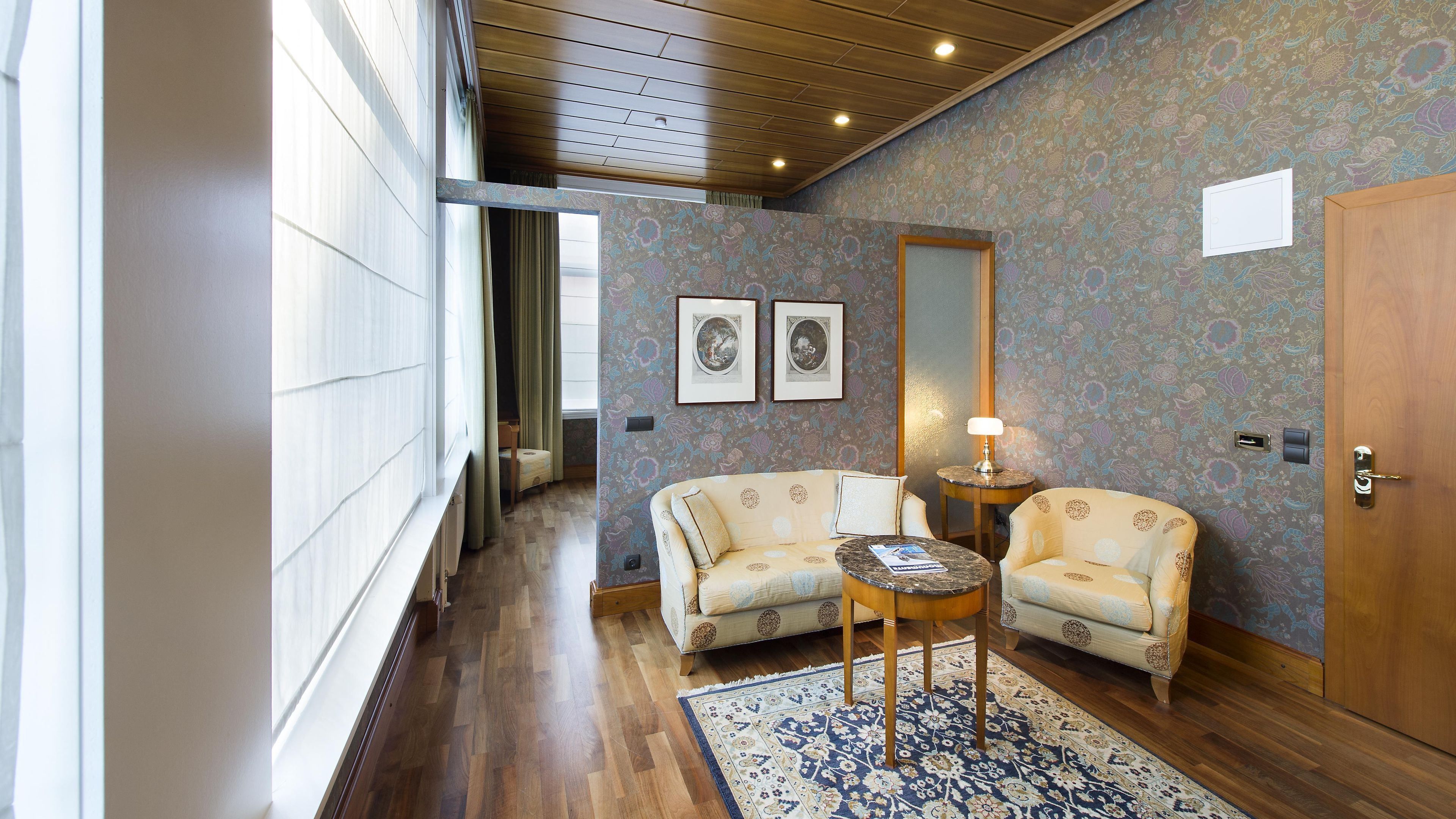 Une chambre d’hôtel avec un plancher en bois, deux fauteuils, une petite table, une lampe, une porte et une fenêtre avec des rideaux.