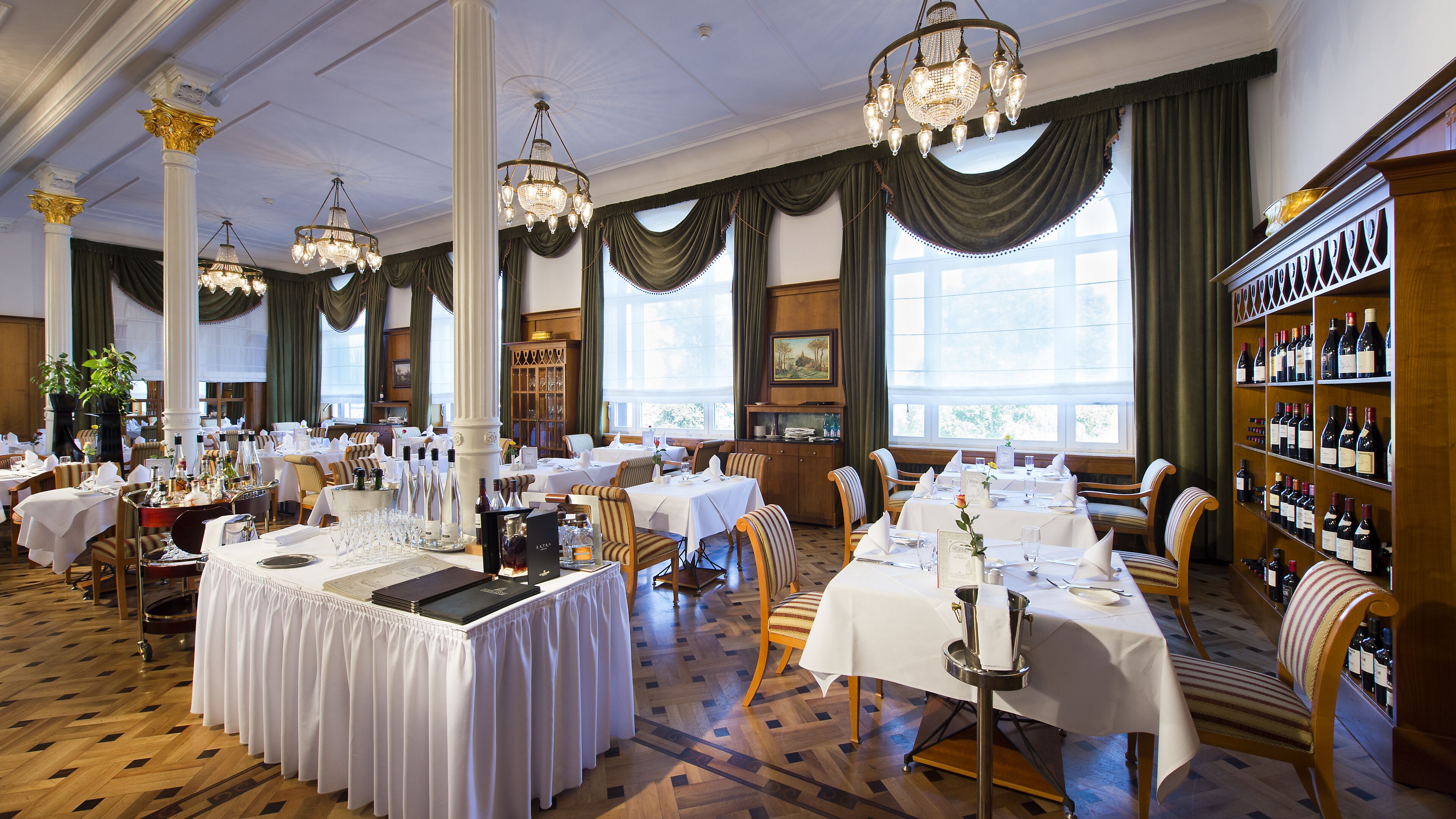 A formal dining room with multiple tables set for service, elegant chandeliers, and a wine rack.