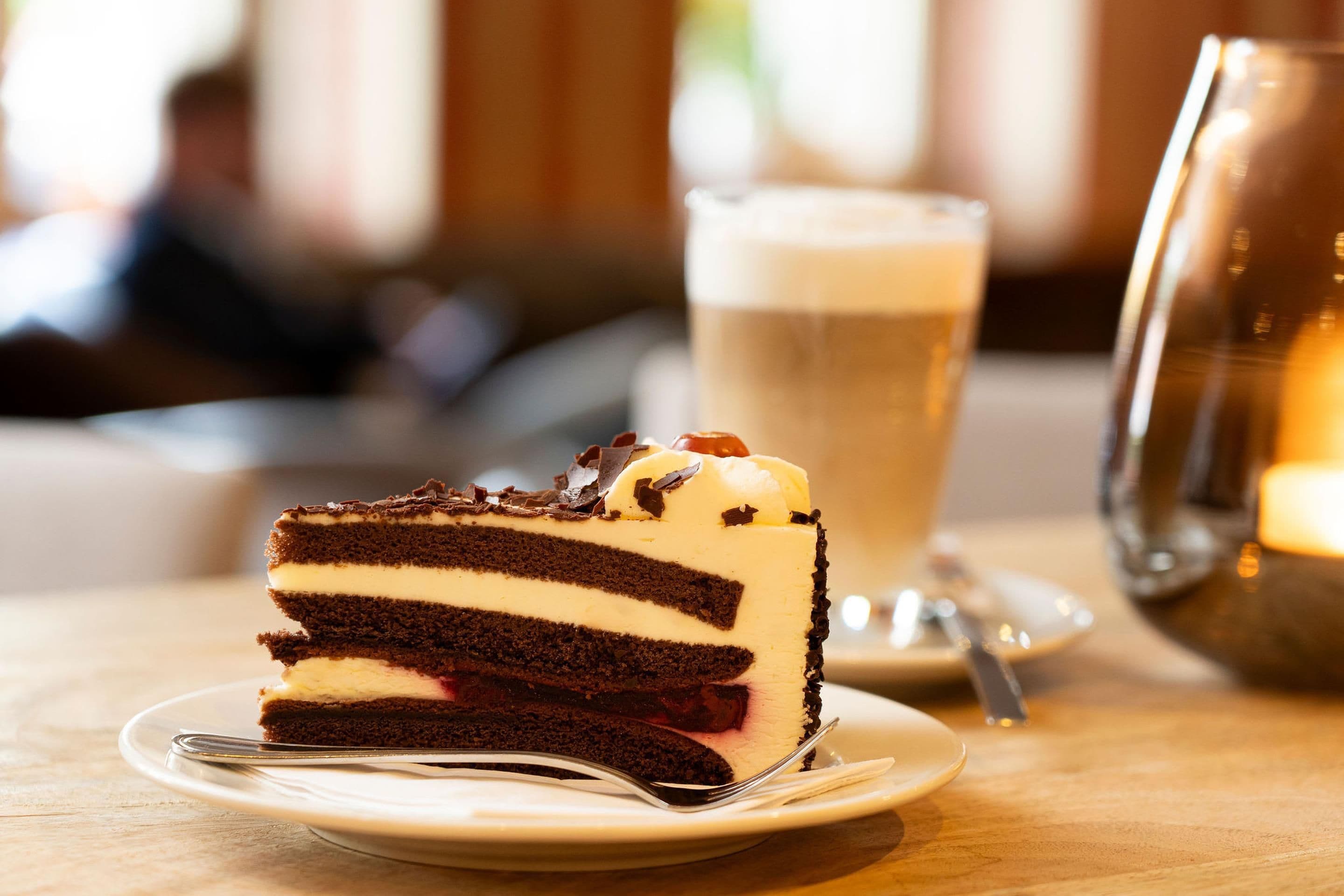 A slice of layered cake with a creamy filling on a plate, a glass of a light-colored beverage, and a metal cup in the background.