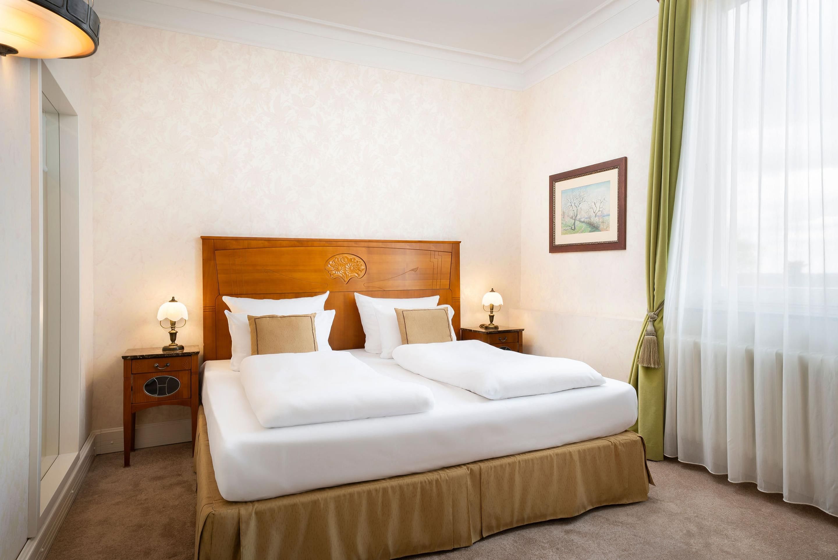 Habitación de hotel con una cama grande, dos mesitas de noche con lámparas, un cuadro enmarcado en la pared y una ventana con cortinas traslúcidas.