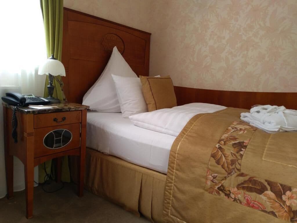 Habitación de hotel con cabecera de madera, cama con ropa blanca, colcha de estampado floral, mesita de noche con lámpara y teléfono.