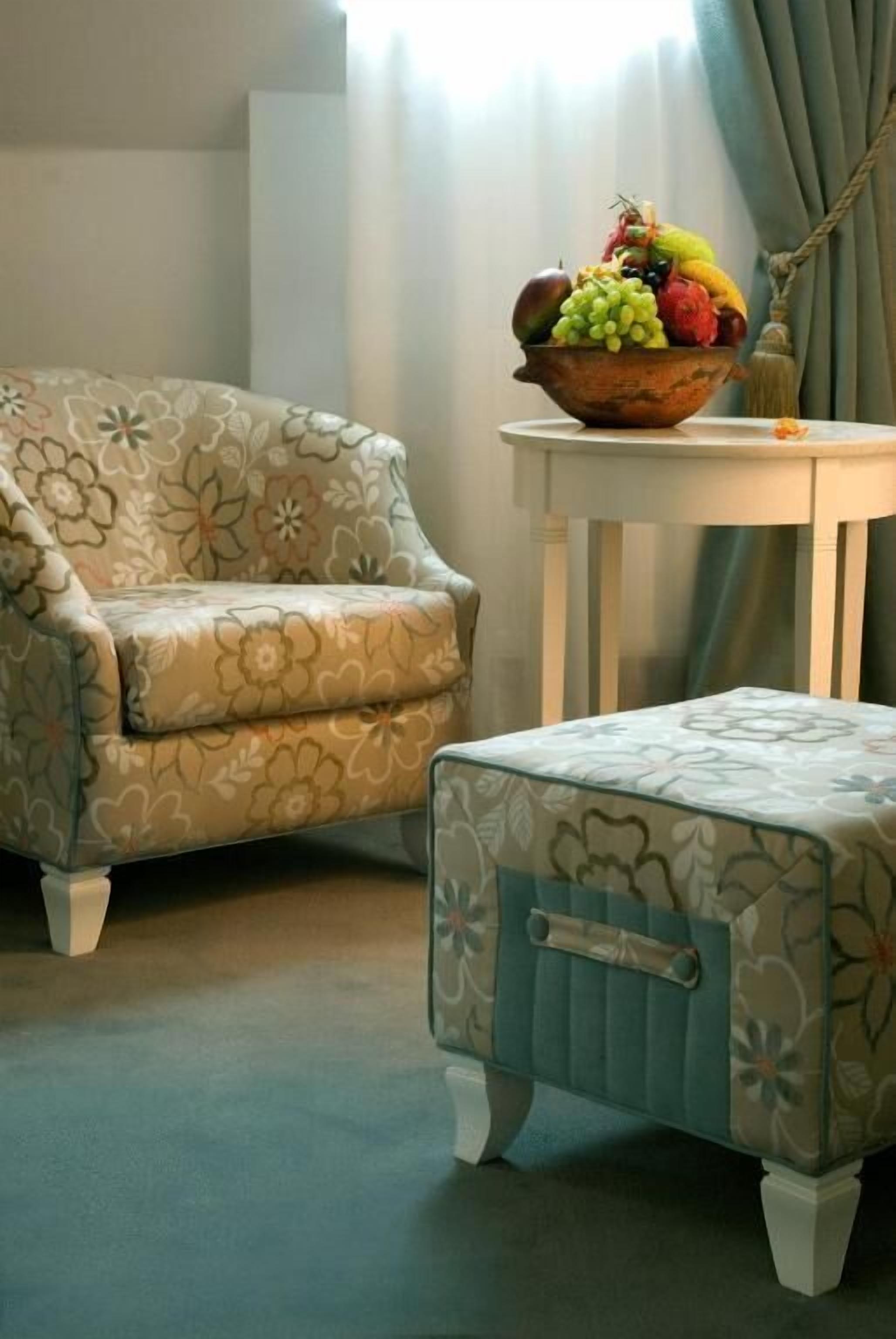 Ein Zimmer mit einer blumengemusterten Couch und Hocker, einem kleinen Tisch mit Obstschale und einem Fenster mit Vorhängen.