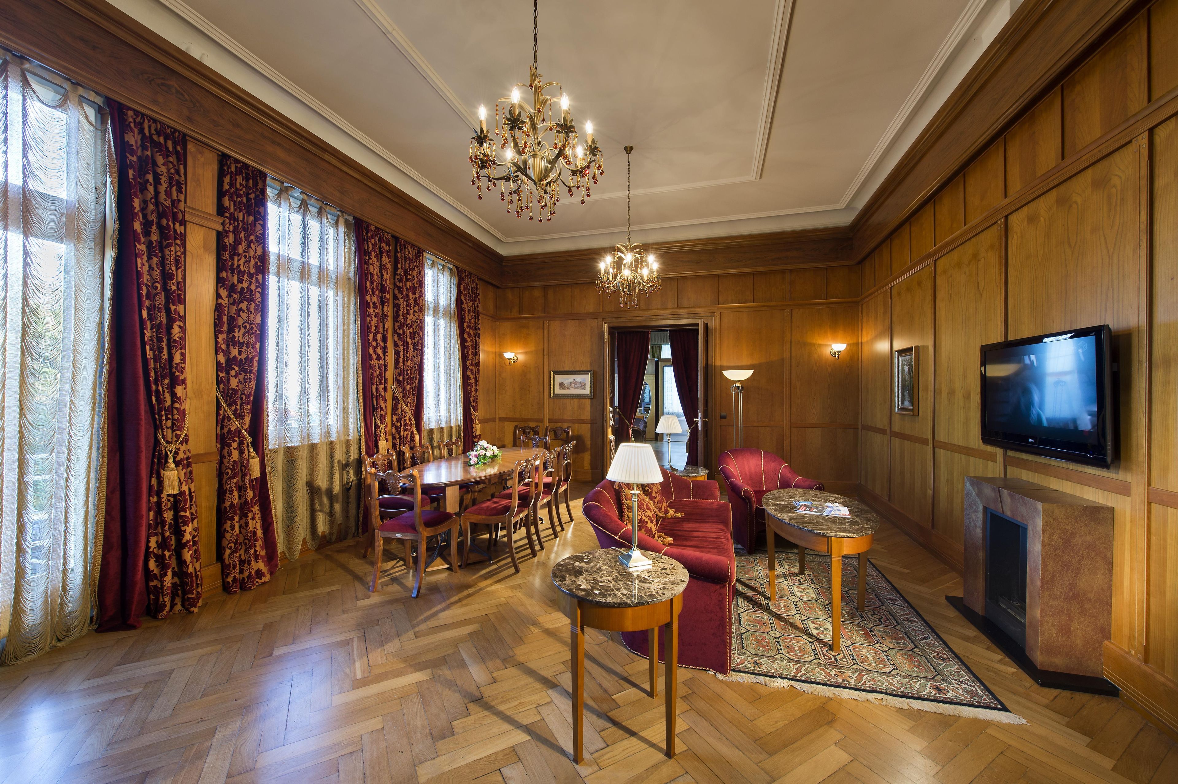 A room with a wooden paneled wall, a chandelier, a dining table with chairs, a sofa, a television, and a fireplace.