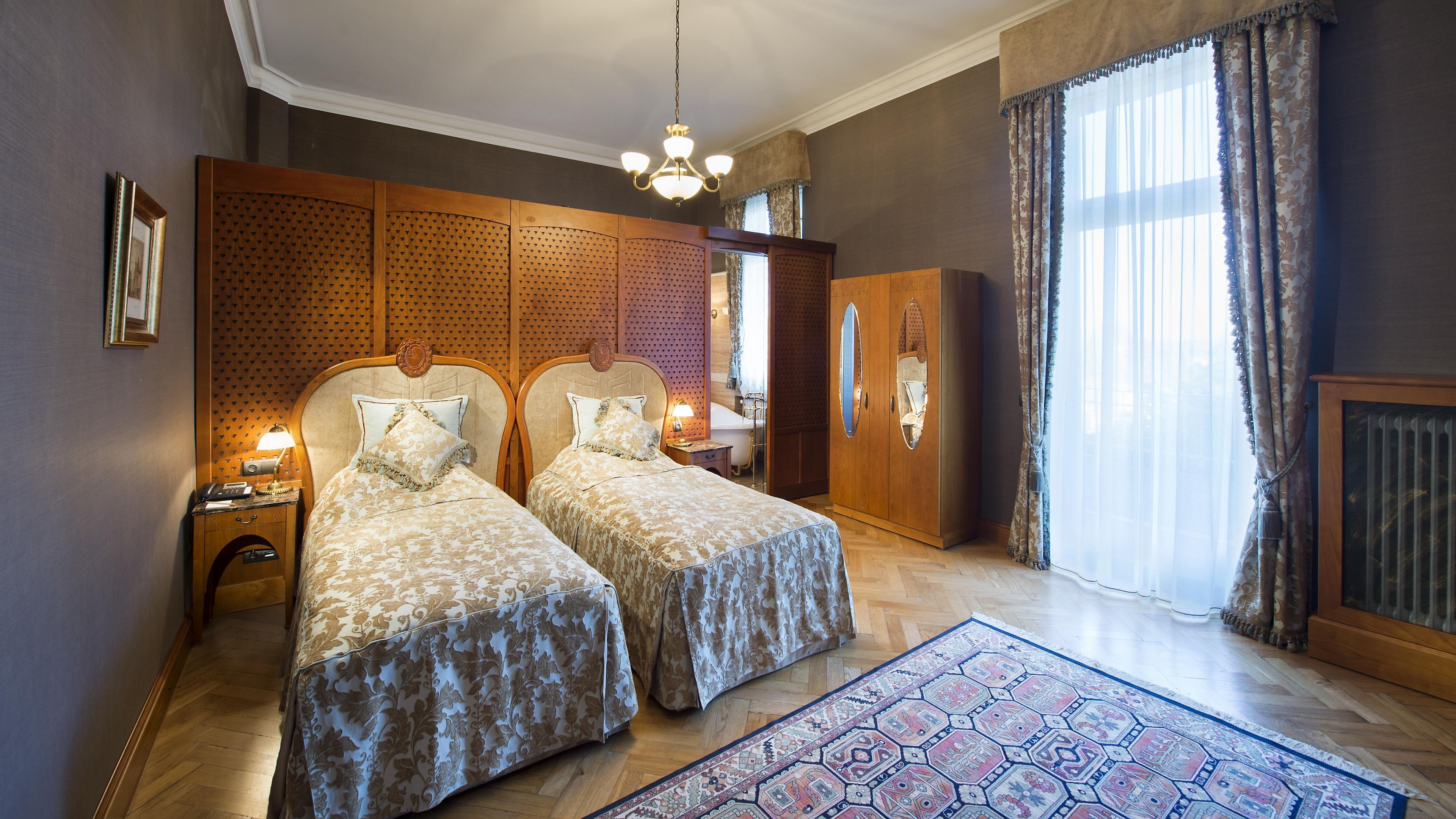 A hotel room with two beds, a wooden wardrobe, a large window with curtains, and a patterned rug.