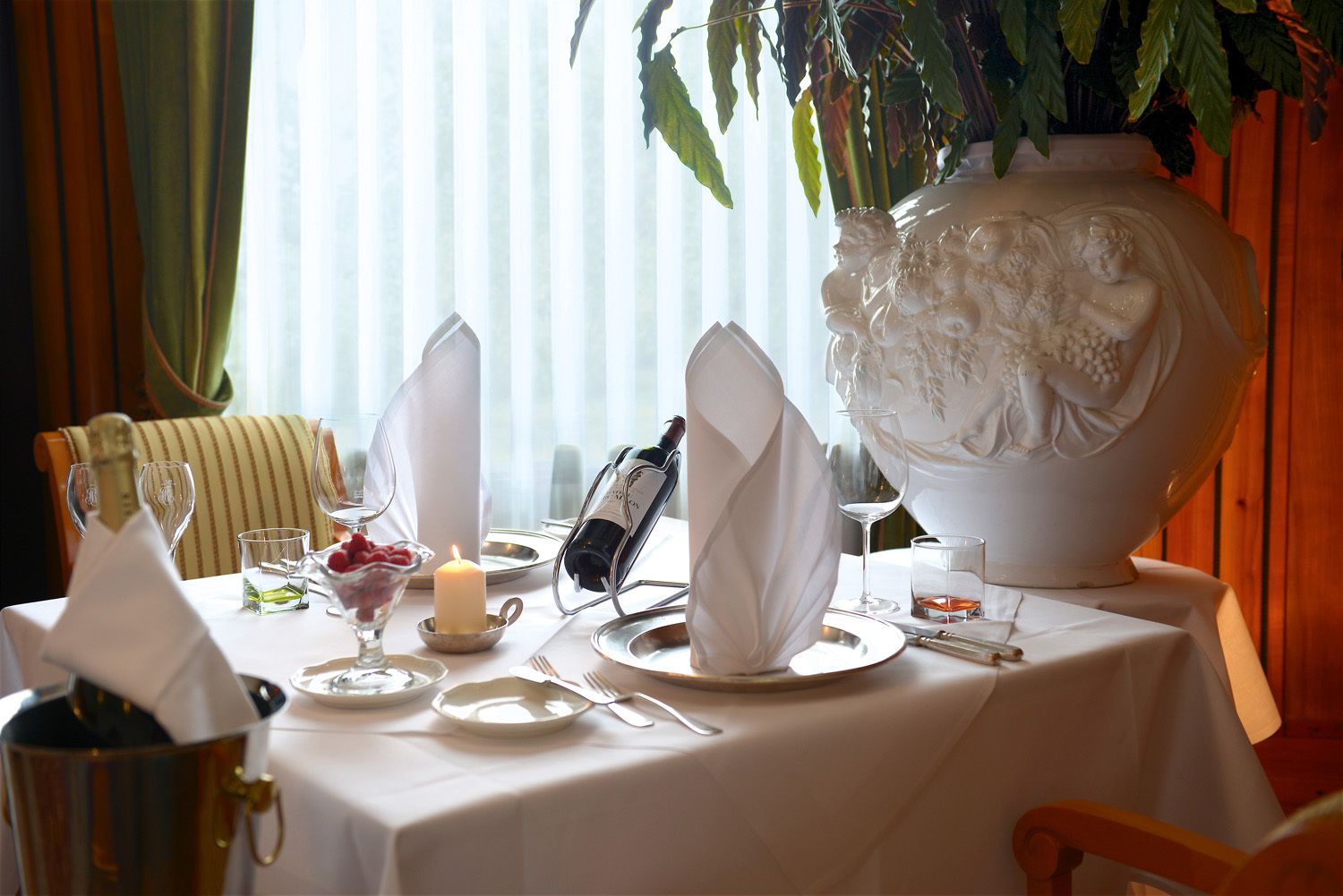 Una mesa lista para una comida, con mantel blanco, platos, vasos, una vela y un gran jarrón decorativo.