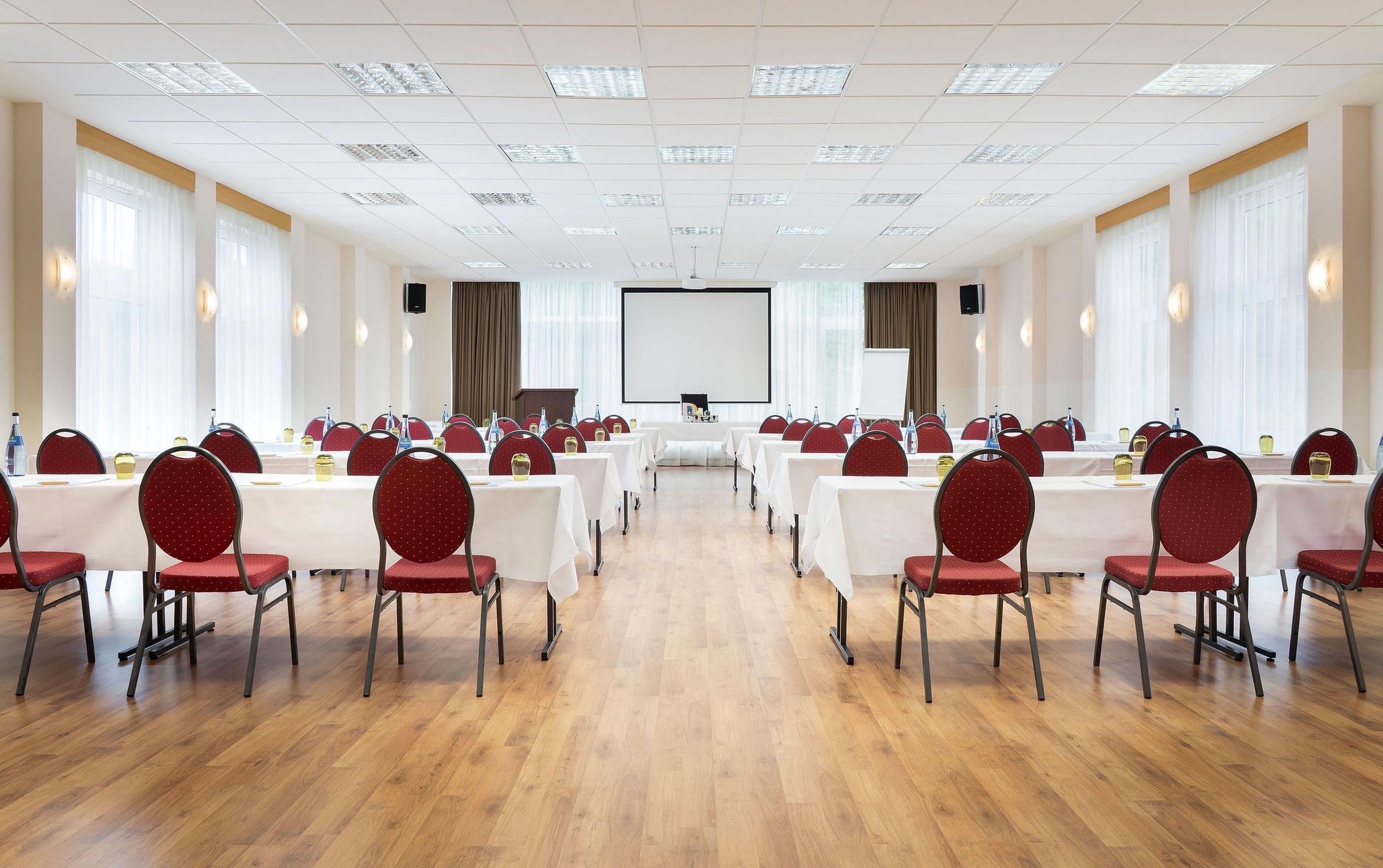 A conference room with rows of tables and chairs, a large screen at the front, and a wooden floor.