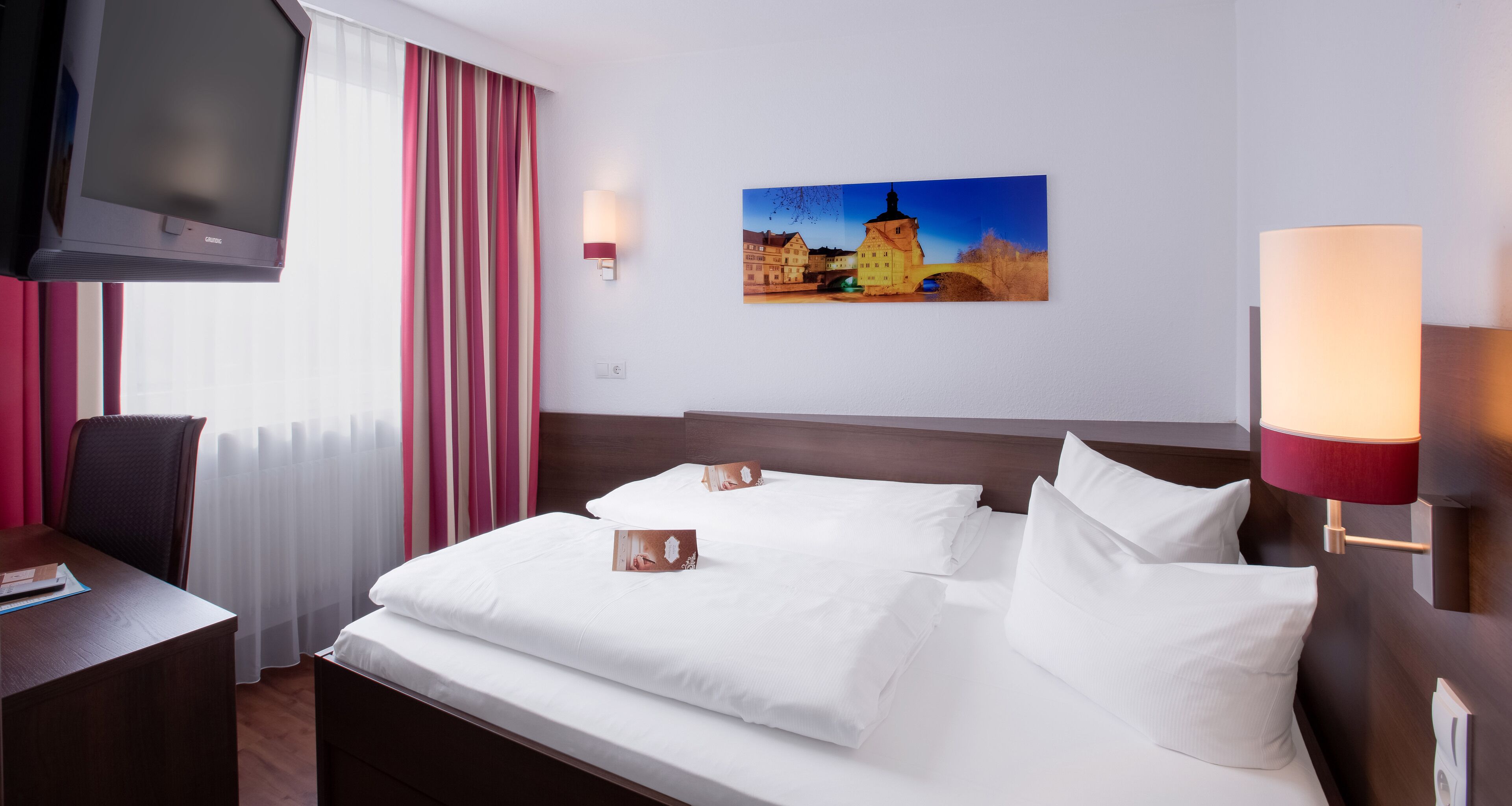A hotel room with a bed, a desk, a chair, a TV, and a painting on the wall.