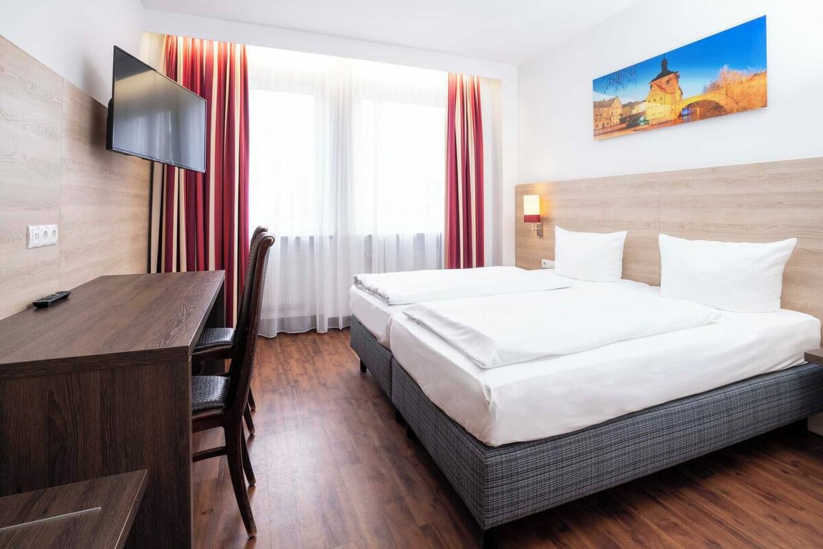 A hotel room with a bed, a desk, a TV, and a painting on the wall.
