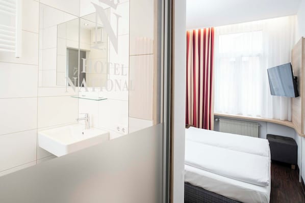 A hotel room with a white sink, a mirror, a bed with white bedding, a TV mounted on the wall, and a window with curtains. - Hotel National (Bamberg)
