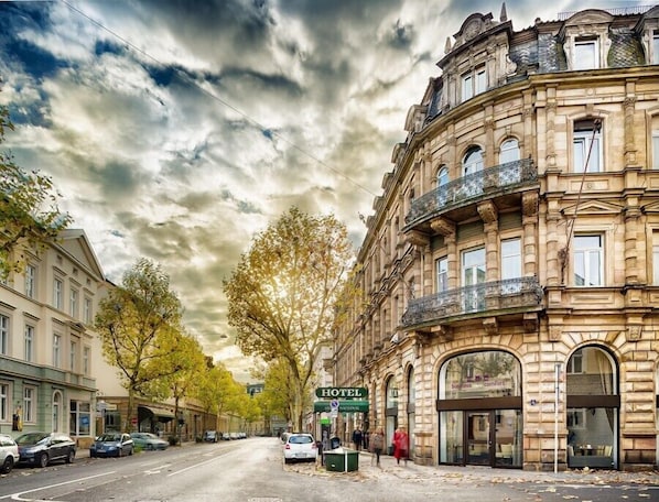 A historic building with a prominent corner entrance, a visible sign for a hotel, and a street lined with parked cars and trees.