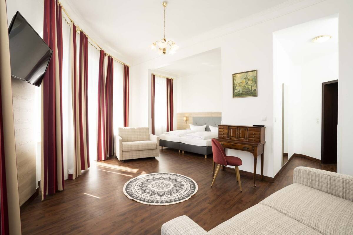 A hotel room with a bed, a desk, a chair, a sofa, a TV, and a painting on the wall.