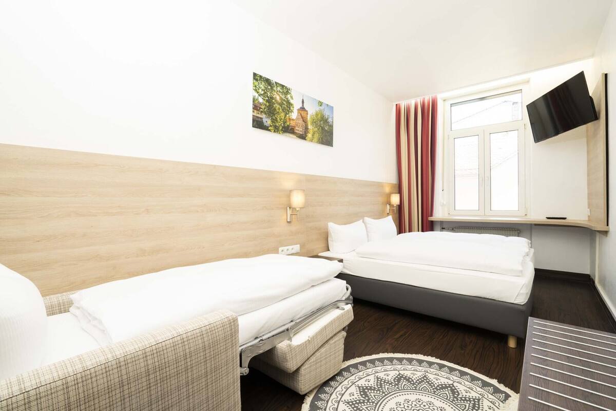 A hotel room with two beds, a flat-screen TV mounted on the wall, a window with curtains, and a patterned rug on the floor.