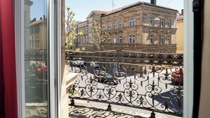 Une vue depuis une fenêtre donnant sur un balcon orné d’une balustrade en fer forgé, un bâtiment à l’architecture classique et un ciel dégagé.