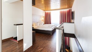 Une chambre d’hôtel moderne avec un plancher en bois, un lit recouvert de draps blancs, un bureau et une vue sur la ville grâce à de grandes fenêtres ornées de rideaux rouges.