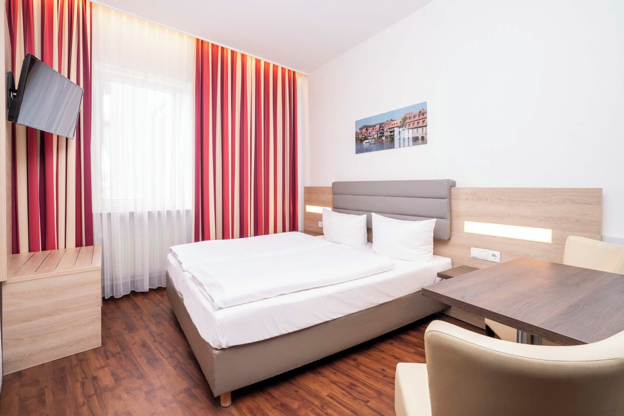 A hotel room with a bed, a desk, a chair, a TV, and a window with curtains.