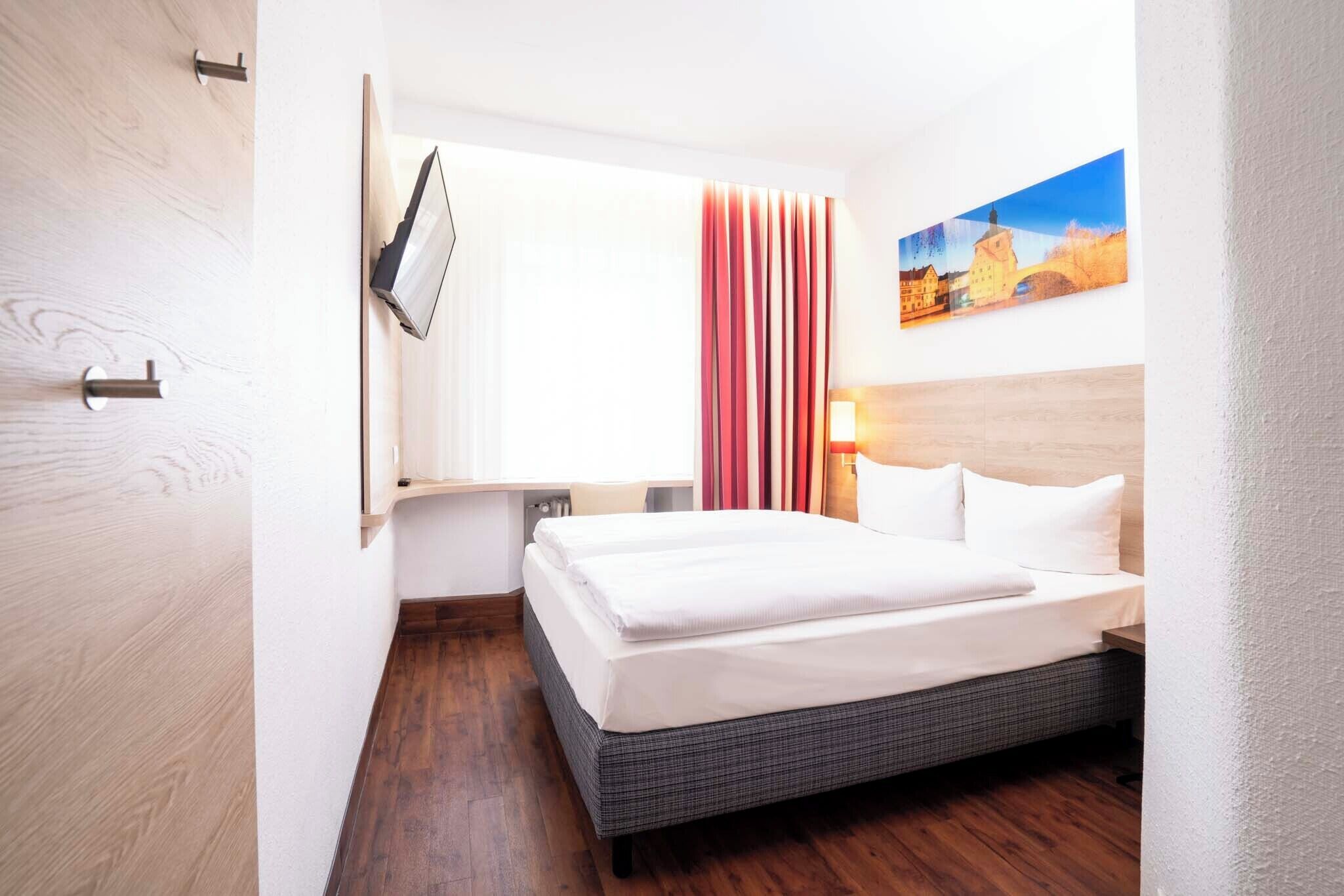 Une chambre d’hôtel avec un lit, un téléviseur fixé au mur, un tableau accroché au mur et une fenêtre avec des rideaux.