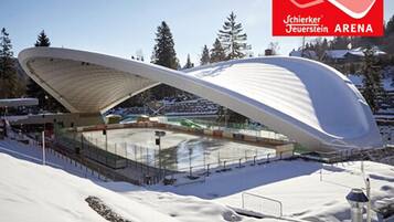 Un estadio moderno, blanco y curvo con techo, rodeado de terreno y árboles cubiertos de nieve.