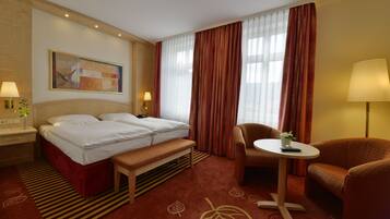 Ein Hotelzimmer mit einem Bett, zwei Nachttischen, einer Bank, einem Sessel, einem kleinen Tisch mit Pflanze und einem großen Fenster mit Vorhängen.