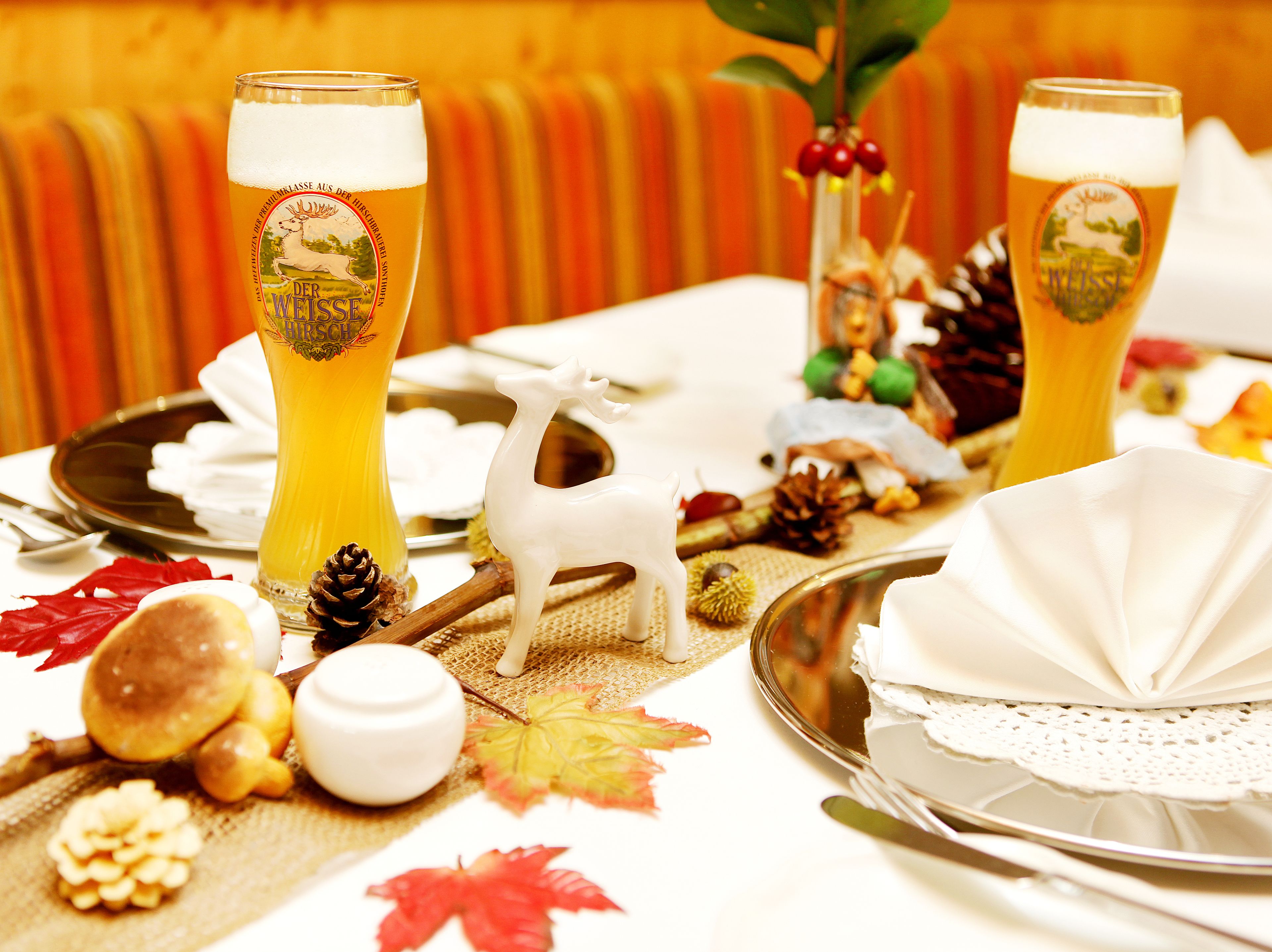 A table set with two tall glasses of beer, a white ceramic goat figurine, pinecones, and autumn leaves.