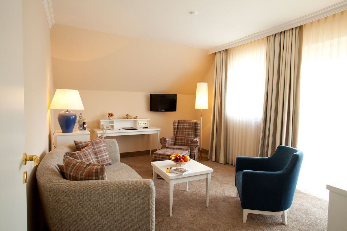 A hotel room with a sofa, two armchairs, a small table, a desk with a TV, and a window with curtains.