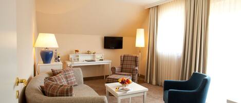 A hotel room with a sofa, two armchairs, a small table, a desk with a TV, and a window with curtains.