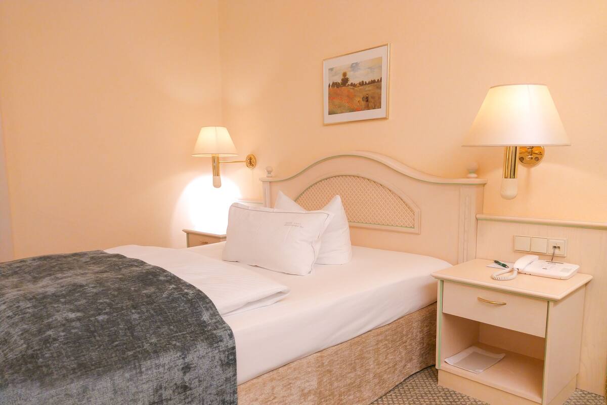 A neatly made bed with white linens and pillows, a bedside table with a lamp, a framed picture on the wall, and a small bedside drawer.