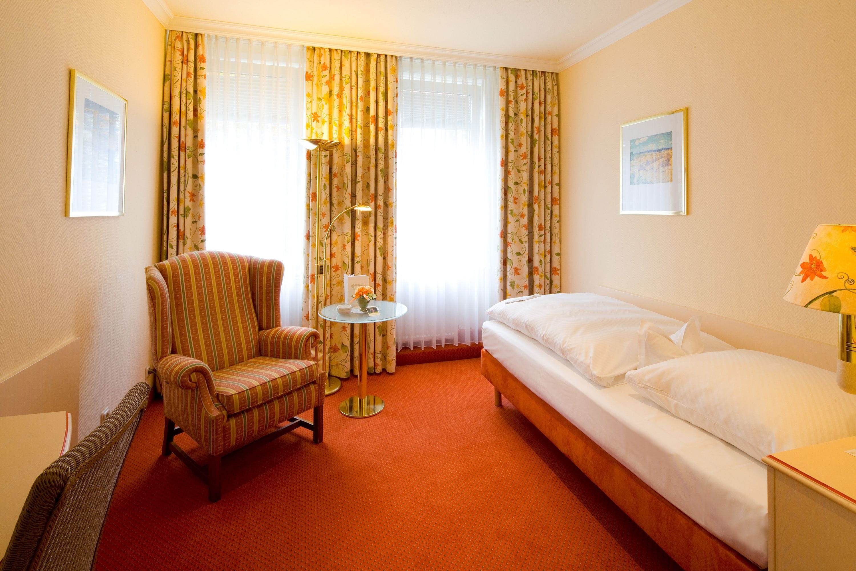 A hotel room with a bed, a chair, a table with a vase of flowers, and two framed pictures on the walls.