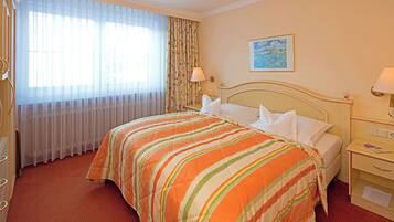 A hotel room with a large bed, a nightstand, a lamp, a painting on the wall, and a window with curtains.