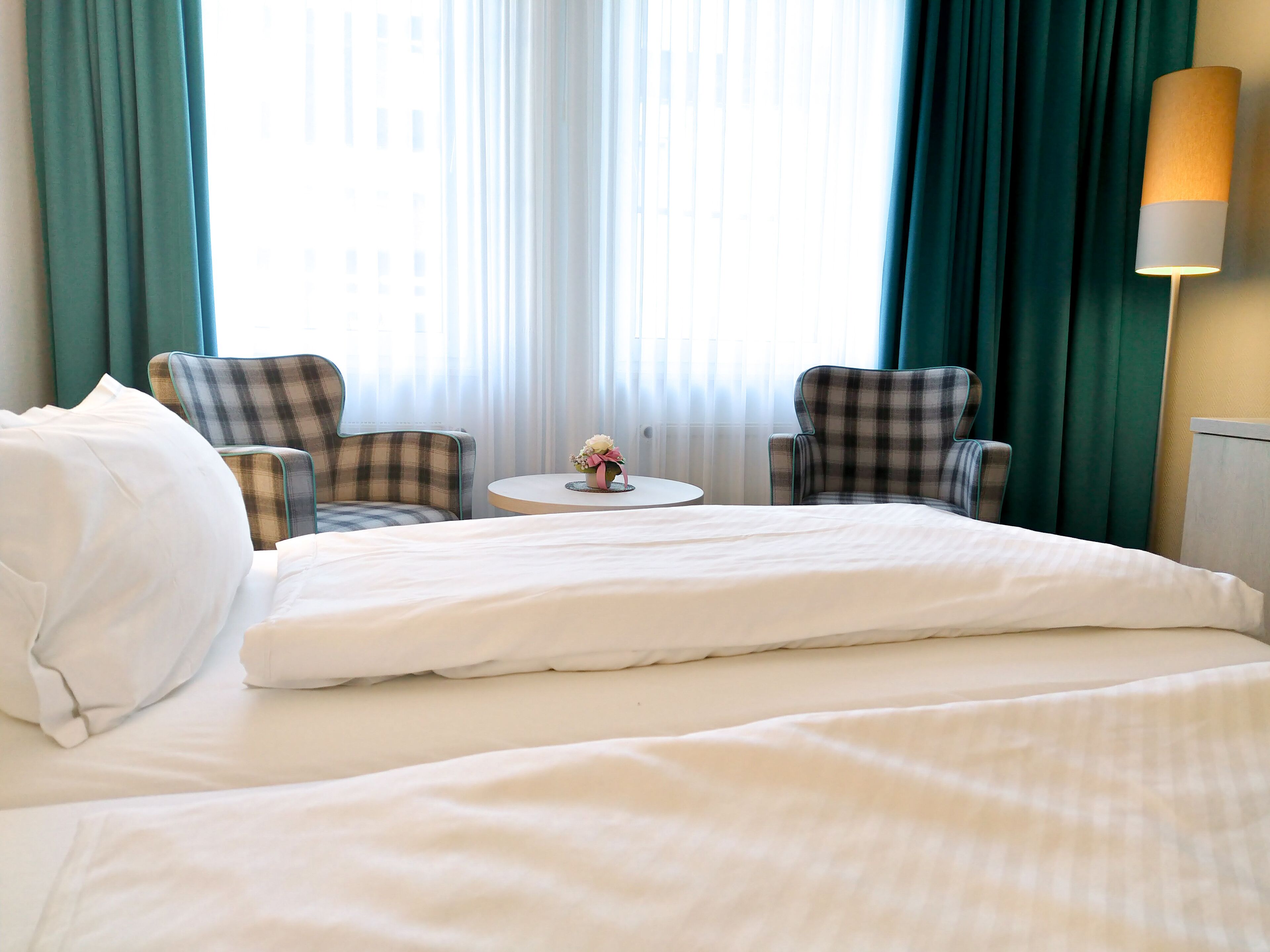 A hotel room with a large bed, two plaid armchairs, a small round table with a vase of flowers, and a lamp on the nightstand.