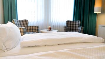 A hotel room with a large bed, two plaid armchairs, a small round table with a vase of flowers, and a lamp on the nightstand.