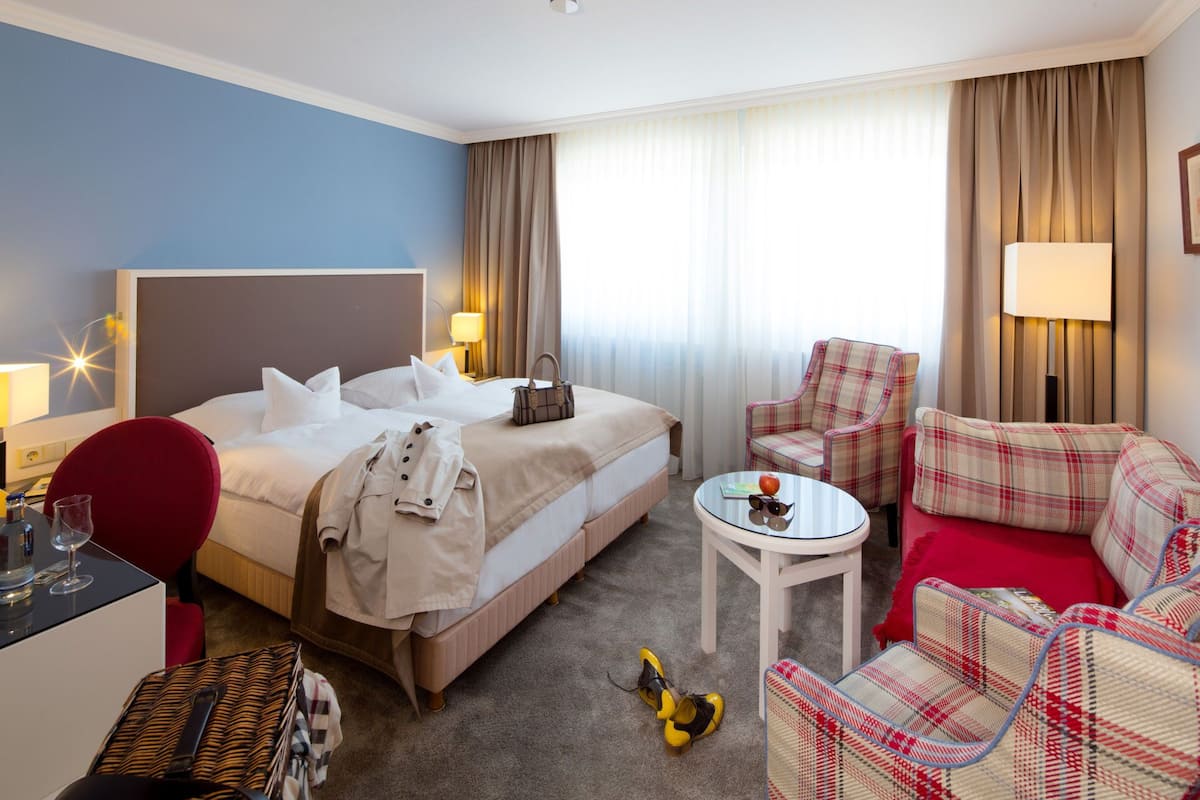 A hotel room with a bed, a desk with a chair, a small table with a lamp, a sofa, and a window with curtains.