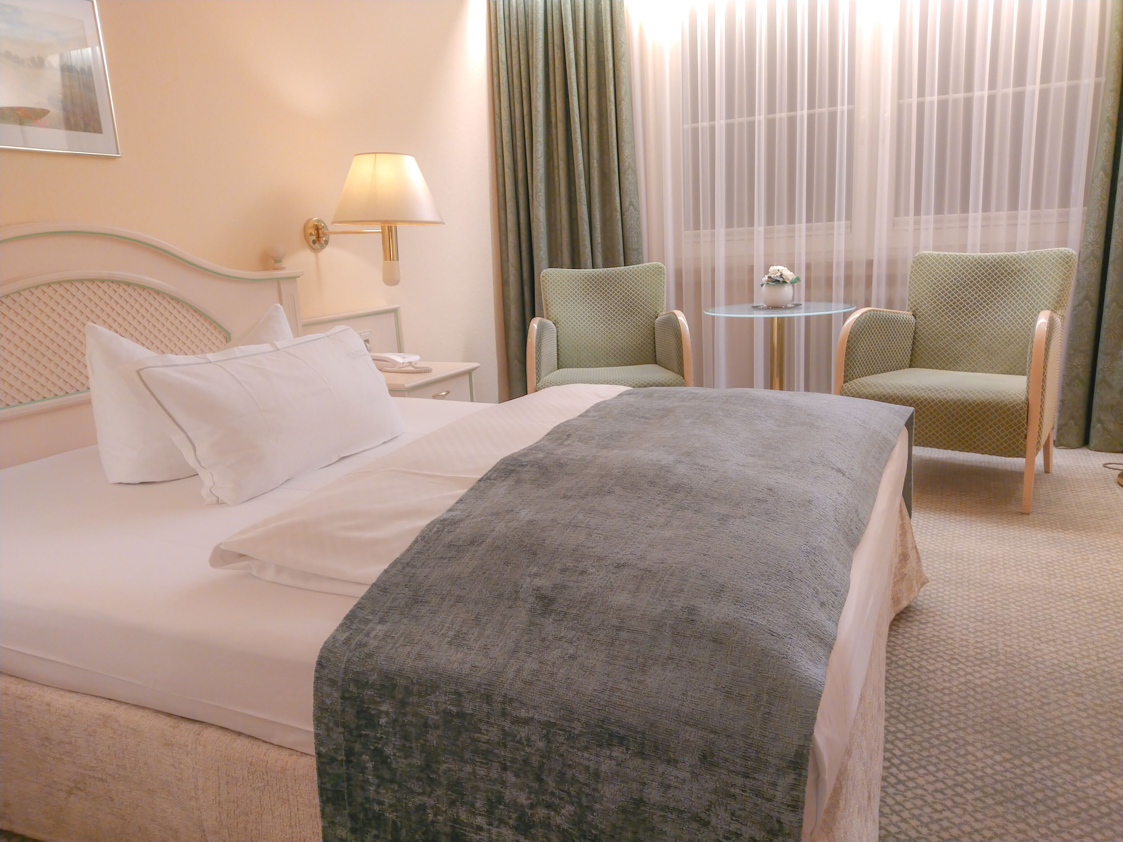 A hotel room with a bed, a bedside table with a lamp, a chair, and a small table with a plant.