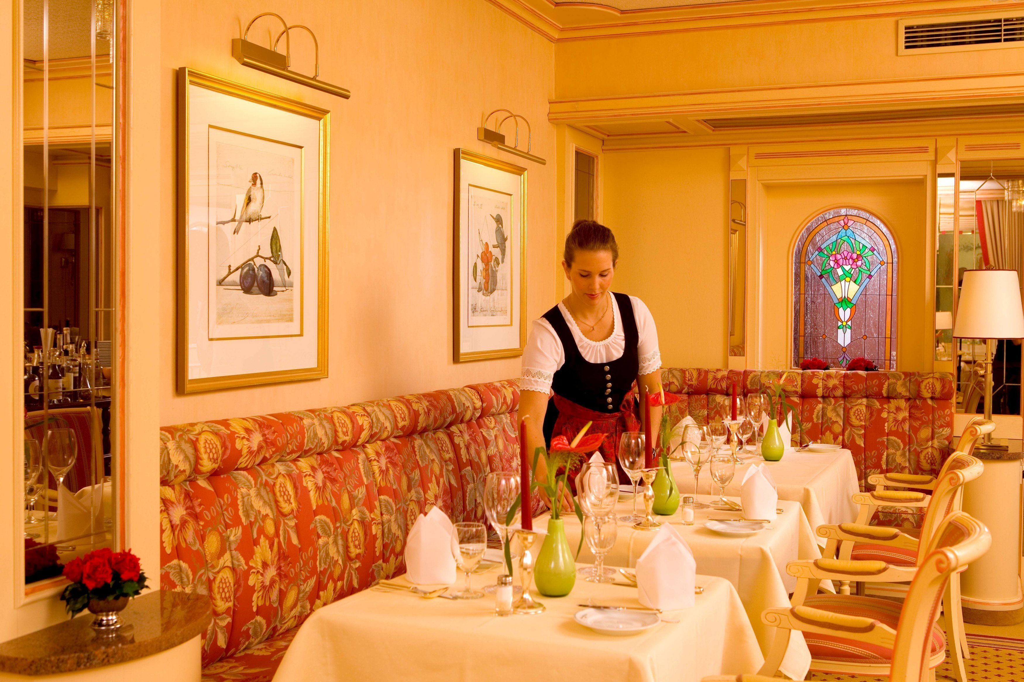 Un área de comedor con un sofá floral rojo, mesas dispuestas con manteles blancos y cuadros en las paredes.