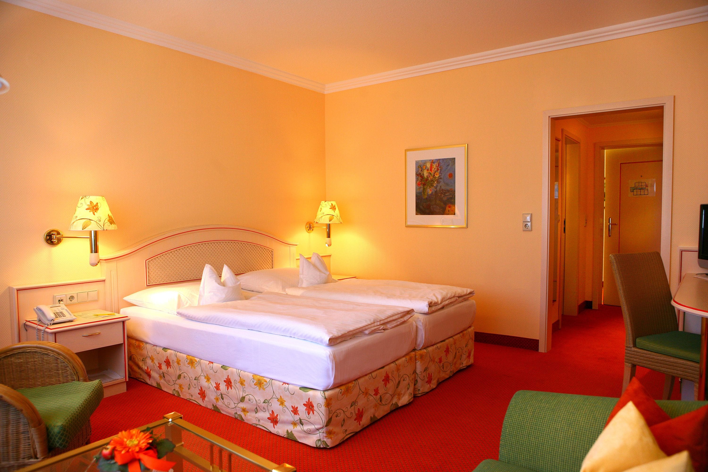 A hotel room with a double bed, two bedside tables with lamps, a chair, a small table with a vase, and a door leading to another room.