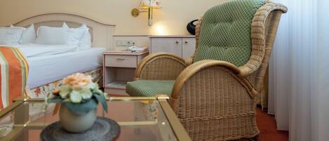 A hotel room with a bed, a green upholstered chair, a bedside table with a lamp, and a small table with a vase of flowers.