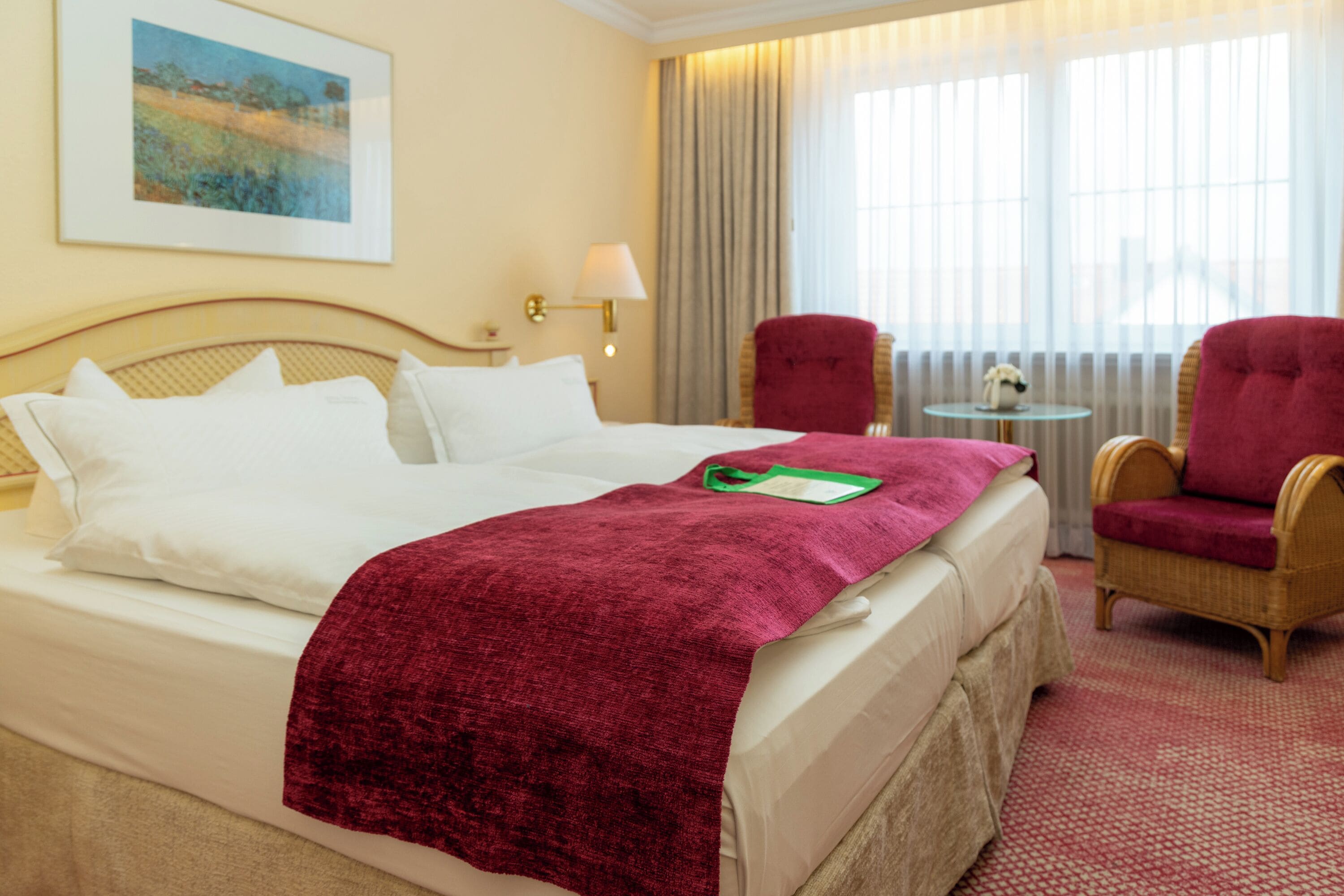 A hotel room with a bed, a red chair, a small table, and a window with curtains.