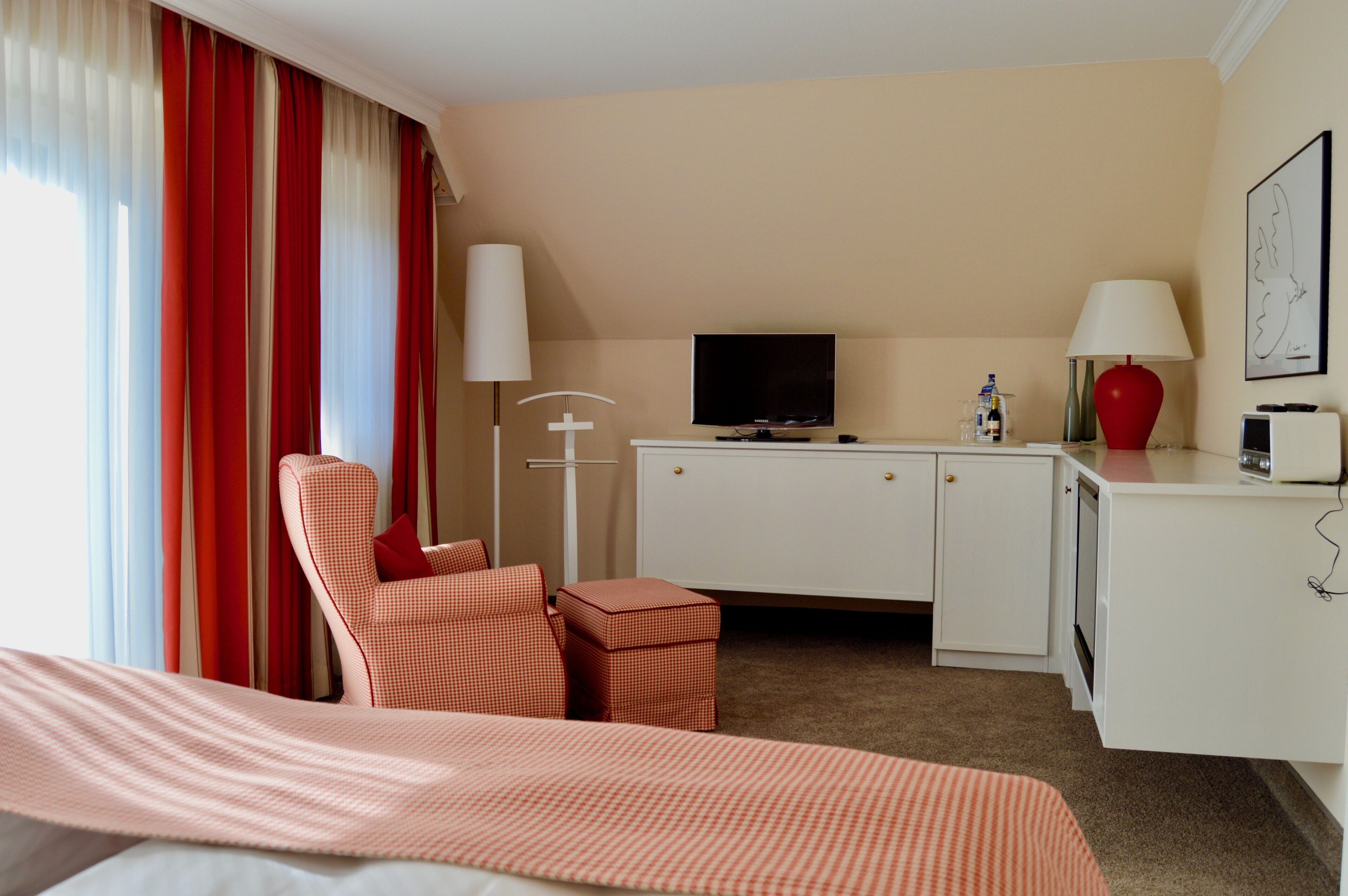 Una habitación de hotel con cama, escritorio con televisor, lámpara roja y silla roja.