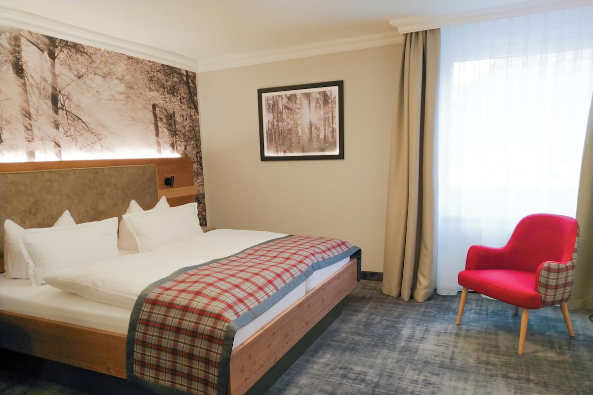 A hotel room with a large bed, a red armchair, a headboard with a light fixture, a framed picture on the wall, and a window with curtains.
